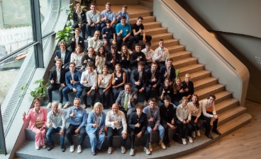 Play-the-Market-Abschlussveranstaltung 2025 Eine Gruppe aus Schüler*innen und Erwachsenen sitzen auf einer Treppe, zeigen den Daumen nach oben und schauen dabei in die Kamera.